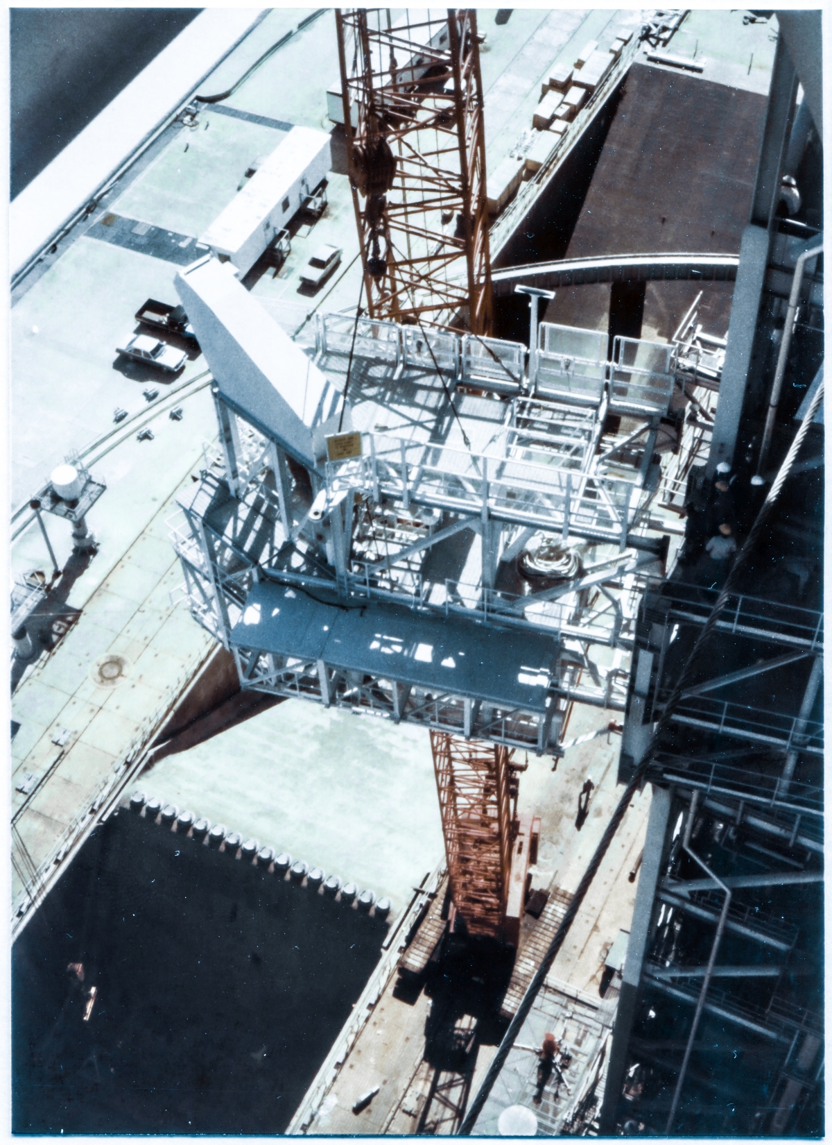 Image 126. Viewed from approximately 75 feet out beyond the north edge of the tower, at the end of the Hammerhead Crane Boom slightly above the elevation 300'-0” Level on top of the Fixed Service Structure at Space Shuttle Launch Complex 39-B, Kennedy Space Center Florida, the Intertank Access Arm can be seen, hanging in free suspension, shortly before it was bolted to its shadowed Support Structure, which you can see personnel standing on, appraising things before the dangerous process of bringing the IAA into direct contact with the steel framing they're standing on top of. Soon, Union Ironworkers from Local 808 working for Ivey Steel will start using a combination of brute force and delicate finesse to align matching sets of bolt holes in the IAA and its Support Structure to a level of accuracy which will permit A325 high-strength structural bolts to be slipped through the matched holes by hand without damaging the threads. Following that, nuts will be placed upon the the bolts, and torqued down, bringing the IAA into firm and final position where it will from then on be able to perform its tasks associated with venting Gaseous Hydrogen from the Space Shuttle's External Tank during fueling operations. Photo by James MacLaren.